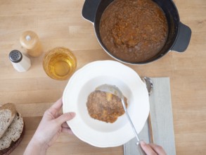 Hands serving lentil stew with ladle from pot to bowl, with bread, water and salt and pepper