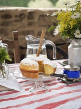 Slice of lemon cake with powdered sugar on a glass stand, pitcher of lemonade, flowers, and