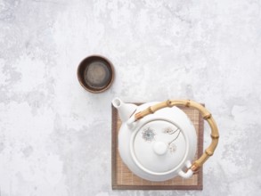 White teapot with bamboo handle and small wooden teacup on bamboo mat, ready for traditional asian