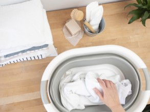 Hand soaking white clothes in basin with natural soap and brush on wooden table, promoting