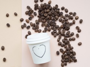 Roasted coffee beans scattered on a two-tone background near a paper cup with a heart drawn on it,