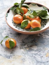 Vibrant tangerines with attached leaves are nestled in a ceramic bowl, placed on a textured, rustic