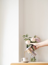 Hand pouring coffee from moka pot into cup on wooden table, with flowers in background, in