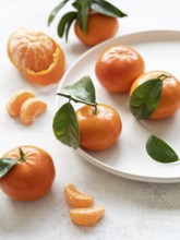 Composition of tangerines with leaves, some peeled and segmented, arranged on a white plate and