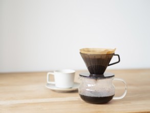 Coffee is brewing using a paper filter and dripper on a wooden table, with a white cup and saucer