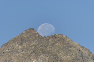 Majestic mountain peak stands tall against vibrant blue sky as large moon rises just behind it. The