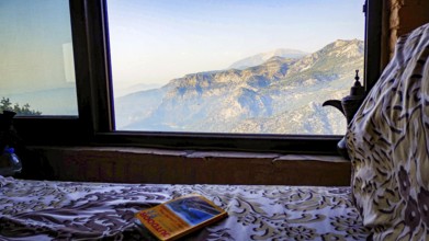 A cozy bed with a book lying on, with a view of a mountainous landscape through a window,