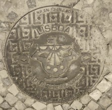 Engraved manhole cover on cobblestones with decorative patterns and a city coat of arms, Lisbon,