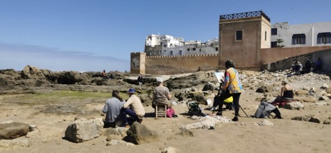 People sitting with canvas on the rocky beach of Essaouira the city view in front of the city wall