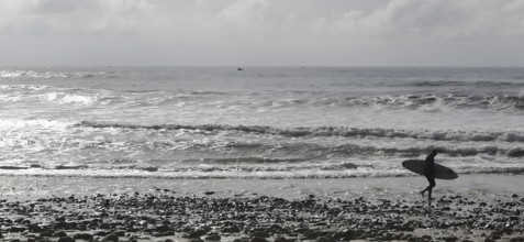 Silhouette of a surfer carrying his board along a pebble beach with a view of the wavy ocean, Sidi
