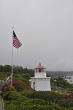 White lighthouse with red roof near the coast, American flag in the foreground, surrounded by