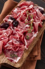 Freshly cut dried meat, served with olives and herbs, on a wooden chopping board, no people