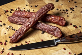 Three salami sticks on wooden board with peppercorns and a knife, crunchy Pfefferbeiser sausage