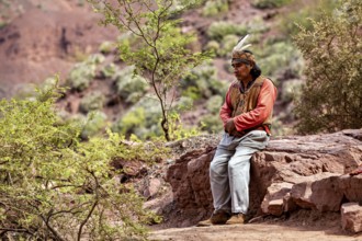 A traditionally dressed man sits thoughtfully on a rock in a desert landscape, a local in the rock