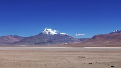 A snow-covered volcano rises above a dry, vast desert under clear skies, the landscape and colorful