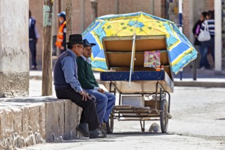 Two men resting under a blue parasol next to a cart, people in the streets of San Antonio de los