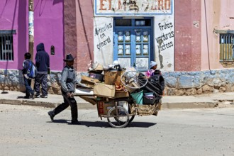Man wearing hat pulls a full cart past a pink building, people in the streets of San Antonio de los