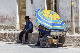 Man and child sitting under a blue parasol next to a cart, people in the streets of San Antonio de