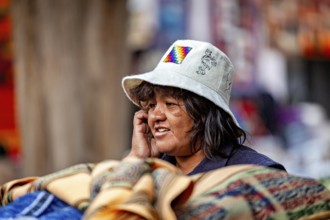 A woman wearing a traditional white hat stands next to colorful fabrics at a market, tourist and