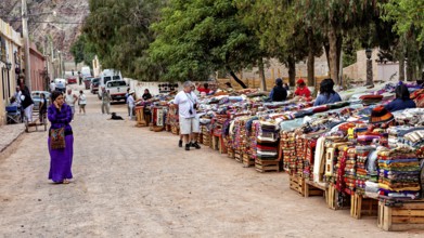 A woman walks along a street with piles of colorful blankets at a market, tourist and souvenir