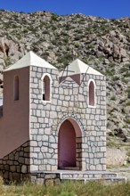Small stone chapel in a remote desert landscape against a rocky backdrop under a blue sky, The