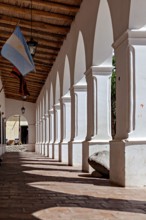 Shady arcade with white columns and wooden ceiling. An Argentinean flag is seen, The arcades of