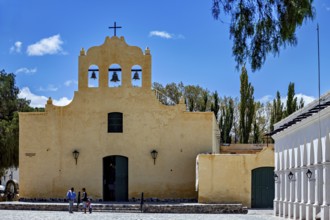 Yellow church with bell tower under blue sky, people on stairs surrounded by trees, Iglesia San