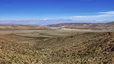 Wide desert landscape under clear blue sky with distant mountains, roads through the Quebrada de