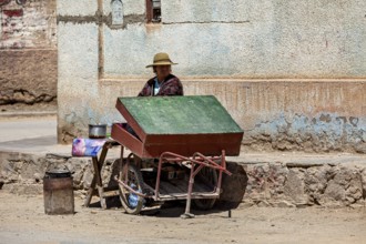 Woman wearing hat sitting at a cart with a green roof on the street corner, people in the streets