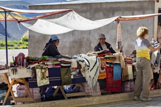 Two woman at market stalls surrounded by handmade blankets, interacting with a customer, tourist