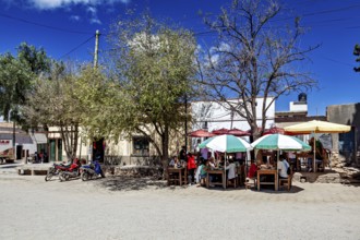 Street scene with people under colorful umbrellas at dining tables surrounded by trees and