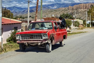 Red, slightly damaged pickup truck on a country road with a man and mountain scenery, old cars in
