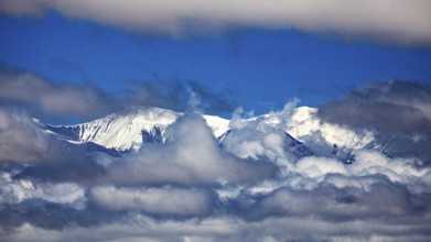 Snow-capped mountains rise through cloud cover under a clear blue sky, The snow-capped peaks of the