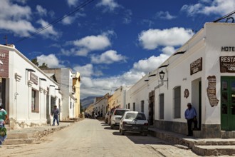 Paved street with traditional buildings, cars on the side of the road and people under a sunny sky