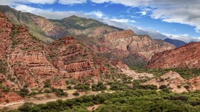 An impressive mountain landscape with red rocks, lush vegetation and a partly cloudy sky, the