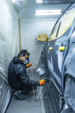 Mechanic in respirator and gloves crouching in a paint booth, spraying a sedan's side panel with a
