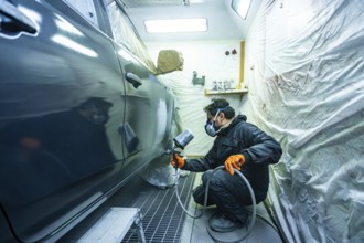 Mechanic wearing respirator mask and orange gloves spraying dark car in a professional paint booth,