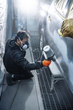 Male auto body mechanic in protective mask and suit applying a fresh coat of gray paint to a car
