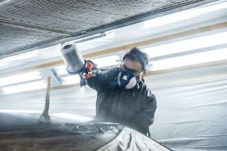 Auto body technician wearing protective respirator and suit painting a car part with a spray gun,