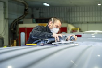 Auto body technician wearing protective mask sanding a vehicle roof with an orbital sander at a