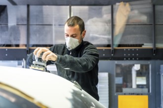 Auto body technician wearing a respirator mask using an orbital sander to prepare a car surface for