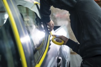 Auto body technician hands applying yellow masking tape around car window trim in a workshop,