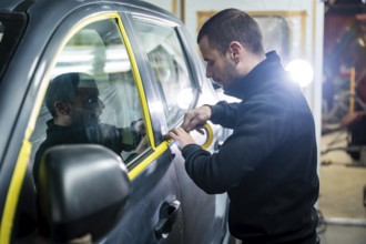 Auto body shop worker carefully applying yellow masking tape to a car door window, preparing the