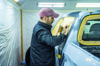 Auto body technician carefully applying masking tape and paper to a car window, preparing the