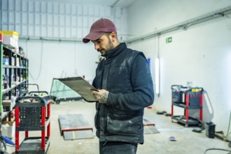 Mechanic in cap and vest inspects vehicle damage in an auto repair workshop, studying a clipboard,