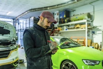 Auto body technician wearing a cap and overalls writing on a checklist, inspecting vehicles in a