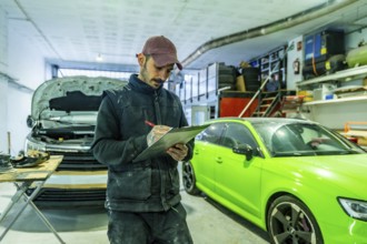 Mechanic with tattooed arms checks inspection checklist on a clipboard in a busy auto body and