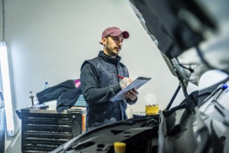Male technician wearing a vest and cap is inspecting a car with the hood open, checking details and
