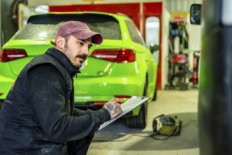 Automotive technician crouching and writing notes on a clipboard during a vehicle inspection or