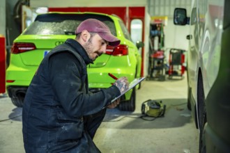 Auto body technician wearing a cap and overalls making notes on a clipboard while inspecting a